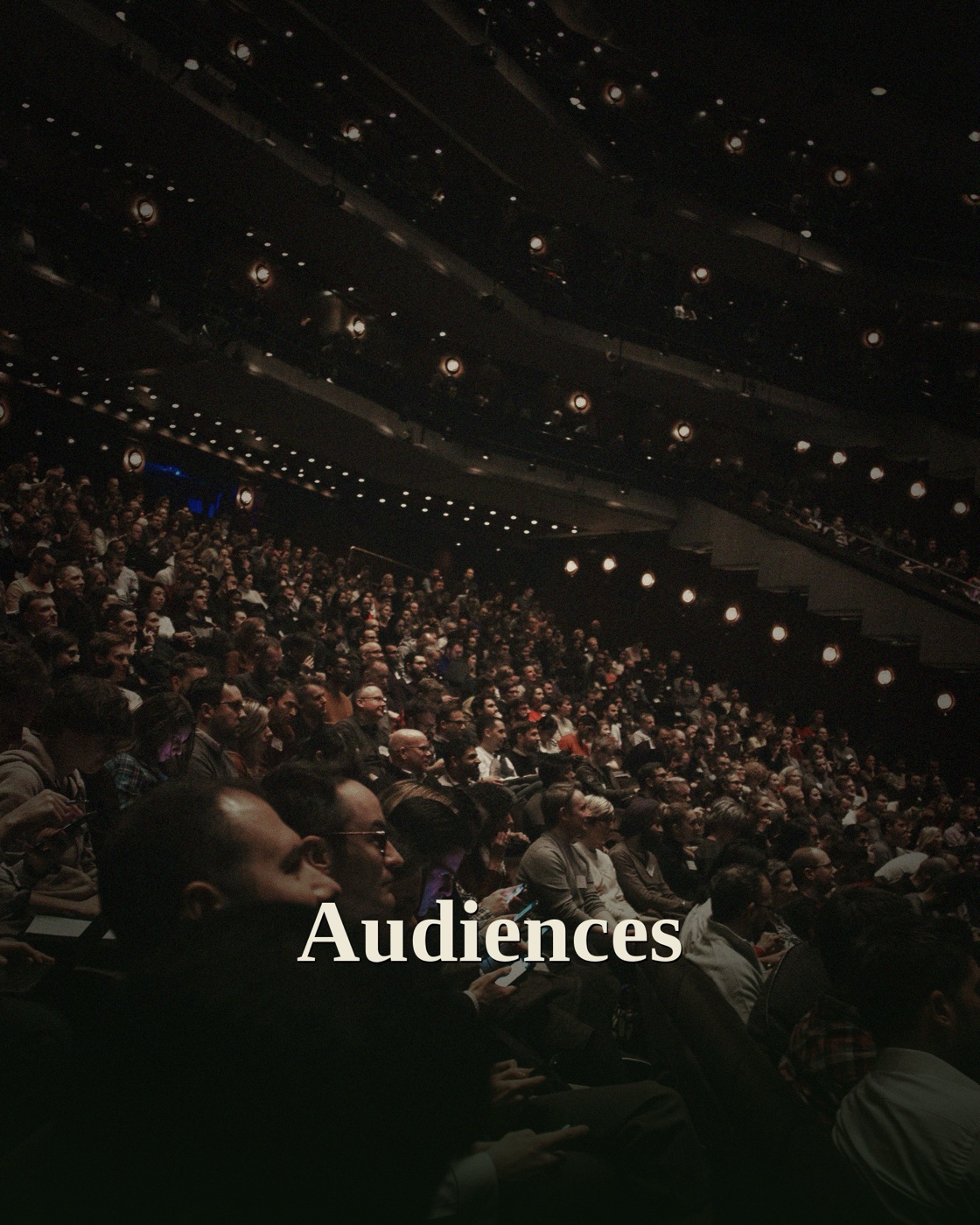 A packed cinema auditorium viewed from the front, dramatic overhead spotlights illuminating a large full crowd