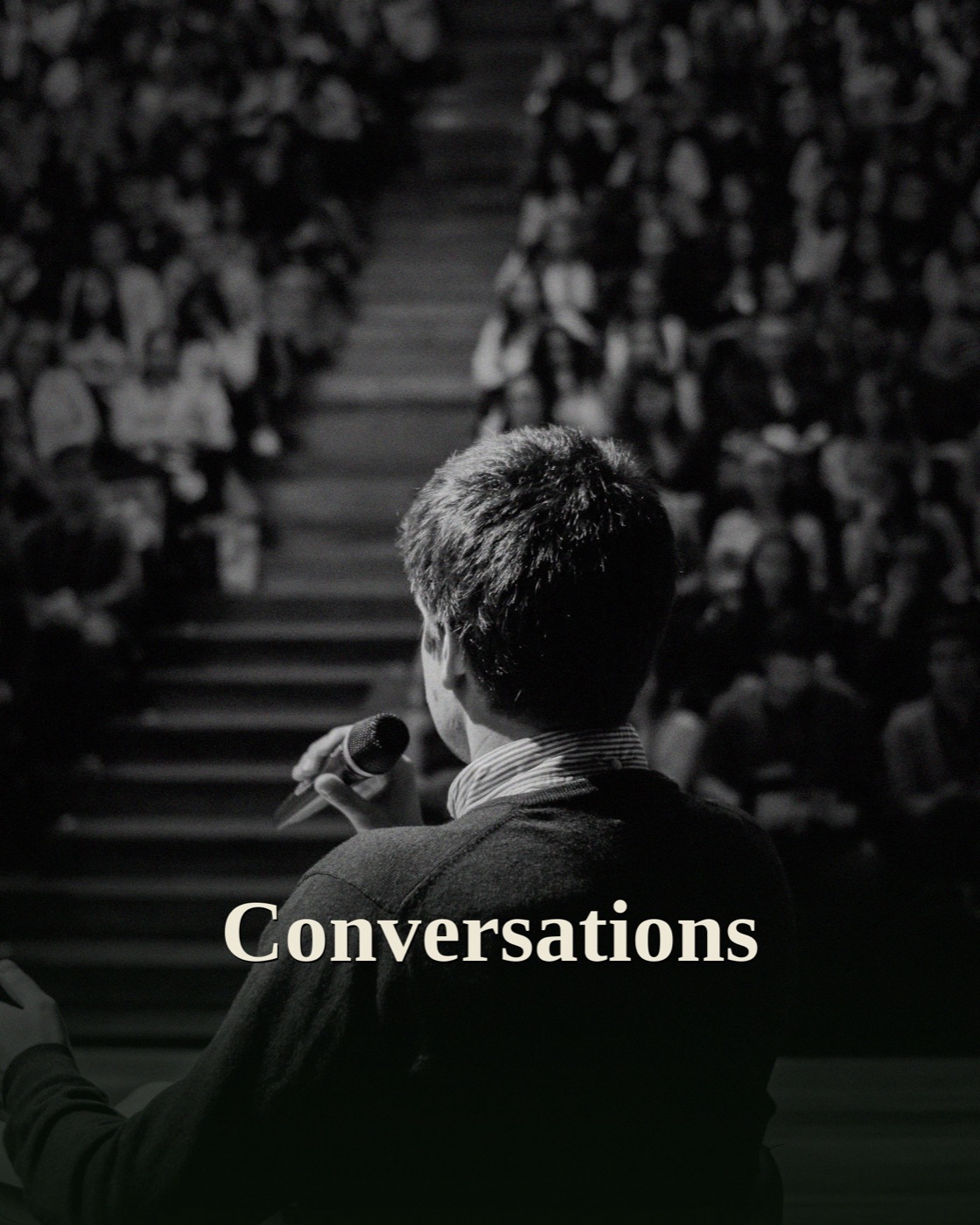 A speaker seen from behind, holding a microphone, addressing a large full auditorium of people, in black and white