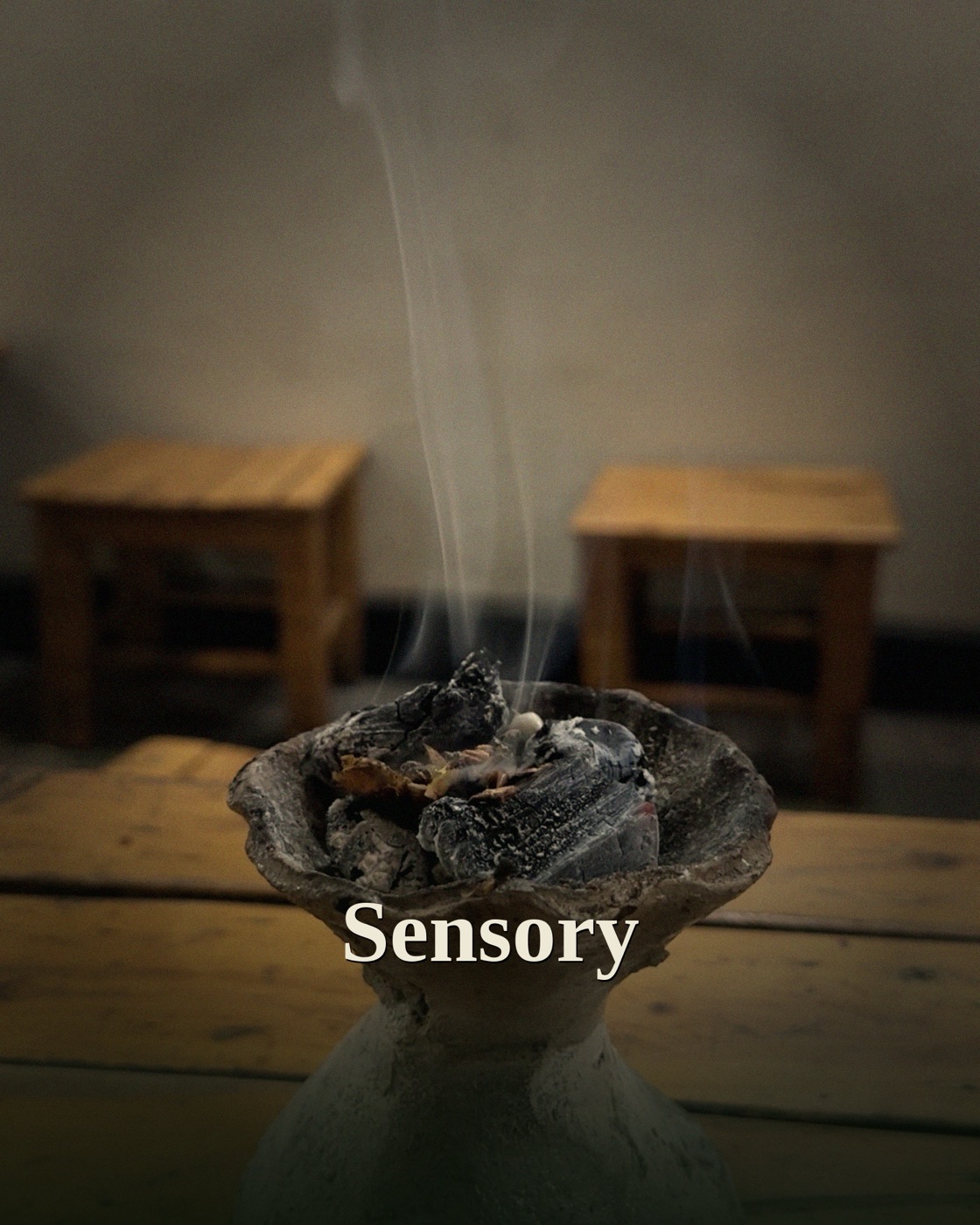 A traditional ceramic brazier with glowing charcoal and a thin thread of smoke rising, set on a worn wooden surface with low stools behind