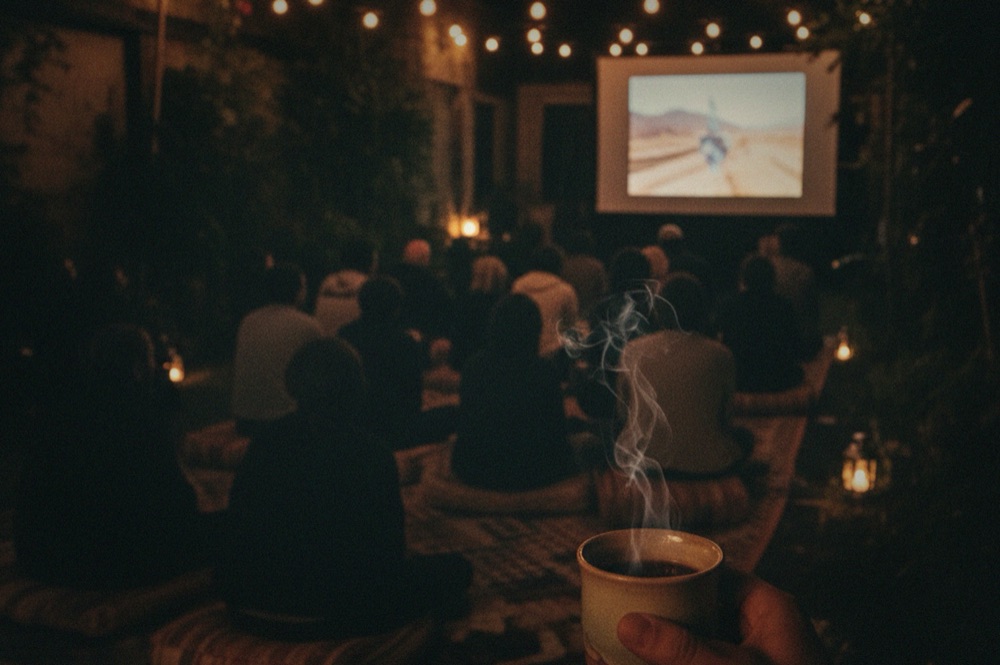 An audience watching a film projected on an outdoor screen at night, warm string lights above, a steaming cup held in the foreground