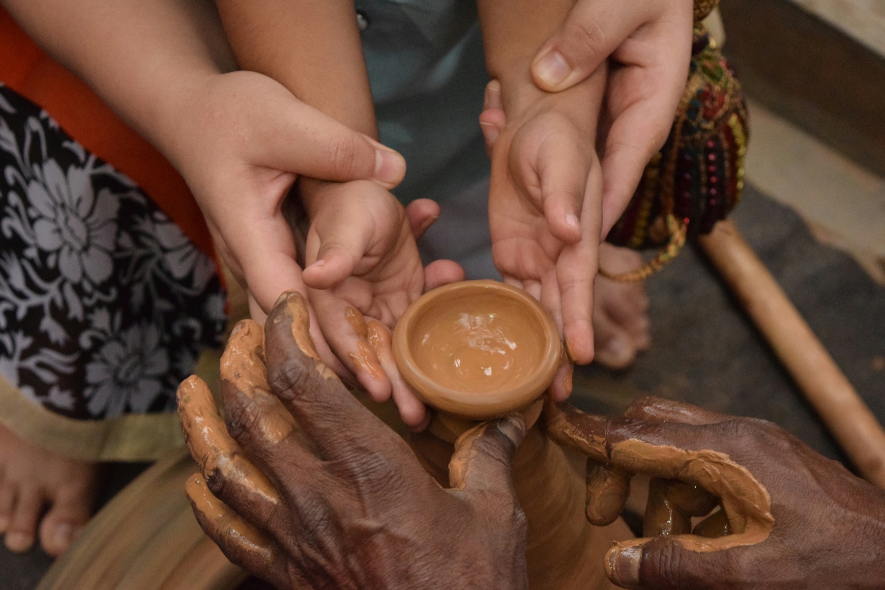 Multiple hands of different ages cradling a small clay bowl, all covered in potters clay