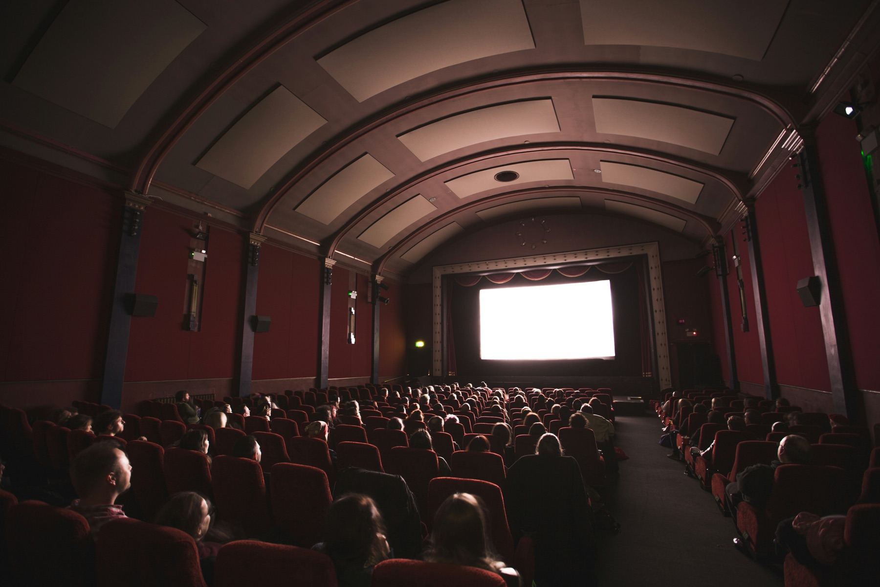 Interior of a classic cinema with red seats, an audience silhouetted against a bright white screen